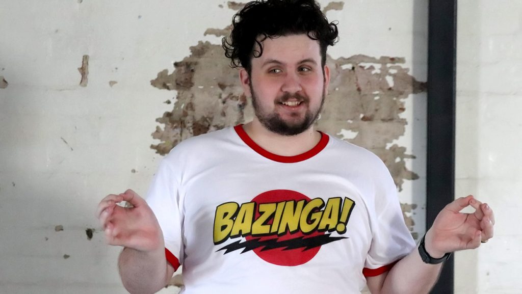 A man standing in front of a brick wall with paint peeling off. He is wearing a white t-shirt with the word Bazinga written across the front