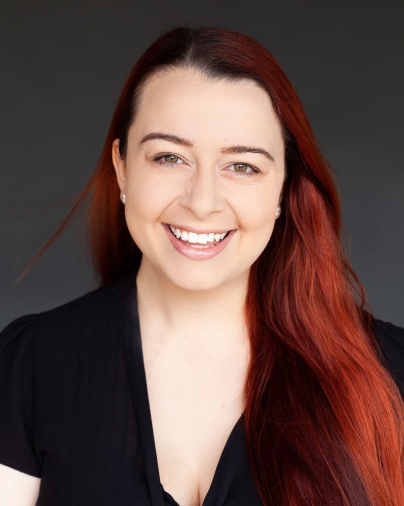 A professional headshot of Madeleine. She has long red hair and is wearing a black top and smiling.