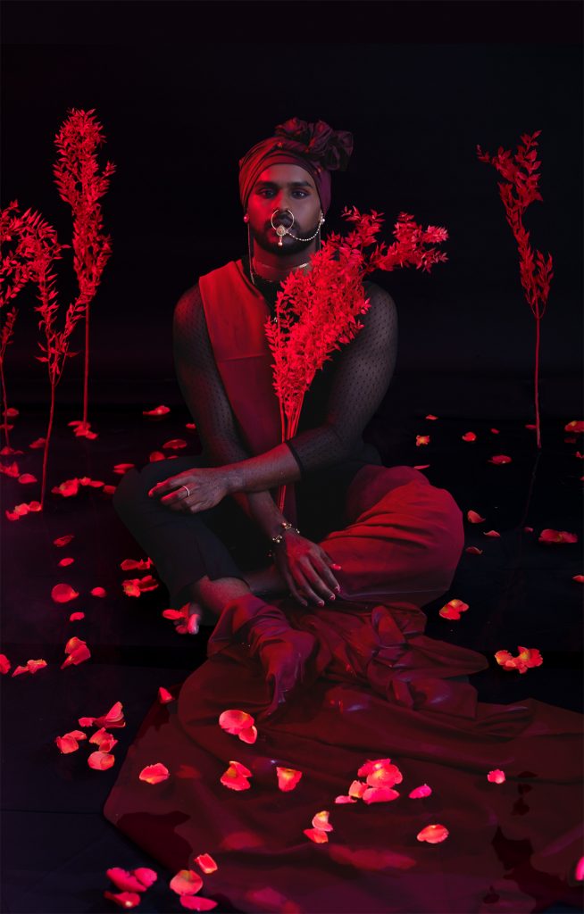An image washed in red. A person sitting cross-legged looks directly at the camera. They are wearing a wrapped head scarf. and have material draped across them. They have a large nose piercing attached by a chain to their ear, and are holding branches of foliage. Petals and branches are in the background.