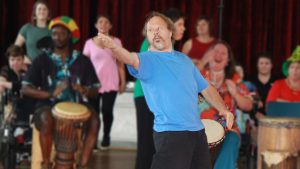 A man standing with one hand held out gesturing to the audience. He is wearing a blue t-shirt and dark pants. There are other performers out of focus in the background.
