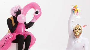 Two figures, one in a white unitard and one in black. The figure in black is draped in an inflatable flamingo, while the white figure is wearing a golden mask.