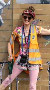A woman standing against a brown brick wall. She is wearing pink tights and a large hi vis vest. Her hair is tied up in a bun on top of her head and she is wearing a hat. One hand is resting on a walking stick, while the other is held out of shot.