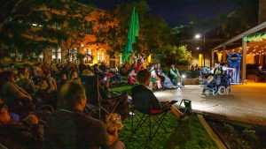 A wide shot of Screech Arts performer Dale on an outdoor stage. He is shown in light, seated in his wheelchair, withe the audience in darkness in front of him.