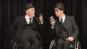 Two men seated in wheelchair wearing black three piece suits and hats. Each is holding a vintage microphone and looking towards one another singing.