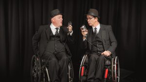 Two men seated in wheelchair wearing black three piece suits and hats. Each is holding a vintage microphone and looking towards one another singing.