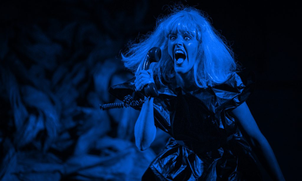 A blue washed image of Andi Snelling mid-performance. She is wearing a blonde wig and holding a phone up to her ear.