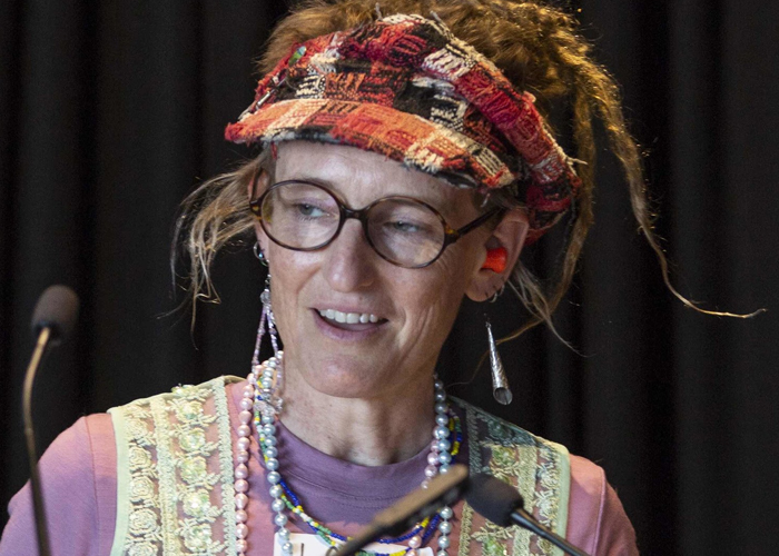 Larissa speaking into two small microphones. She is wearing a tartan visor and reading glasses. Her hair is styled in dreadlocks worn high on her head.