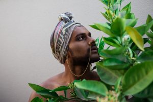 Naavikaran pictured in focus behind green foliage. They are wearing a patterned head scarf wrapped around their head and have a gold chain linking a nose ring to their ear.