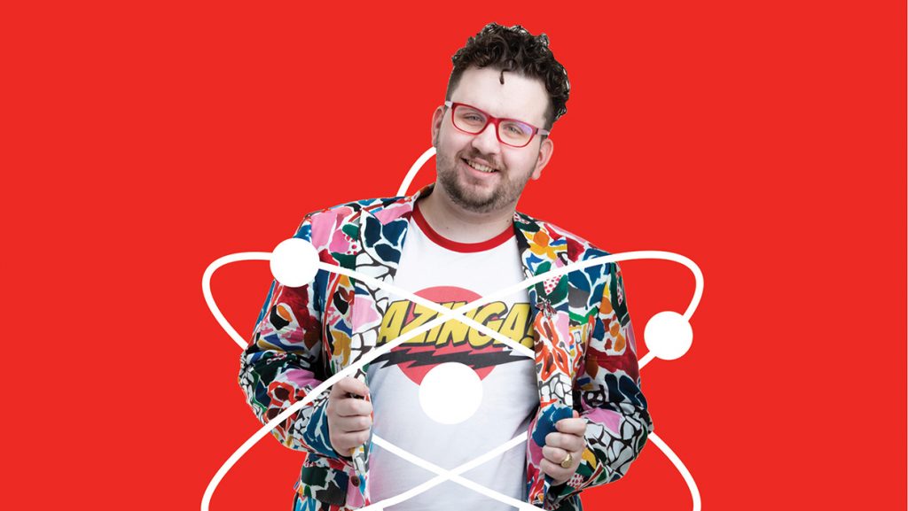 A red background with Oliver standing in the centre wearing a brightly coloured jacket over a t-shirt with the word 'Bazinga' written across it.