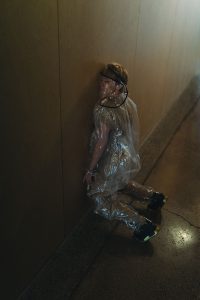 Ruby wearing a see-through plastic outfit and mask. She is leaning her cheek against a wall in an empty hallway.