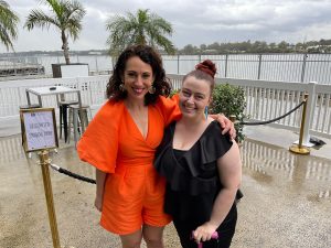 Two people standing close together for a photo outside. Louise Bezzina (left) is wearinga bright orange jumpsuit and smiling. Madeleine Little (right) is wearing a black top and pants and has one hand resting on a pink cane. They are both smiling.