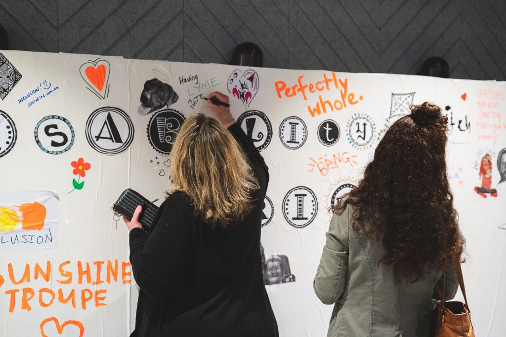 Two females standing with their back to camera, one has brown curly hair, the other with blonde hair. Writing on a white wall filled with test, symbols and icons.