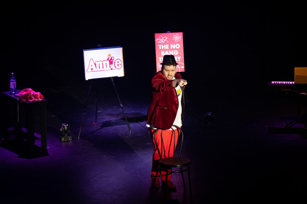 A man on a stage in red pants and jacket with a black hat pointing at an audience.