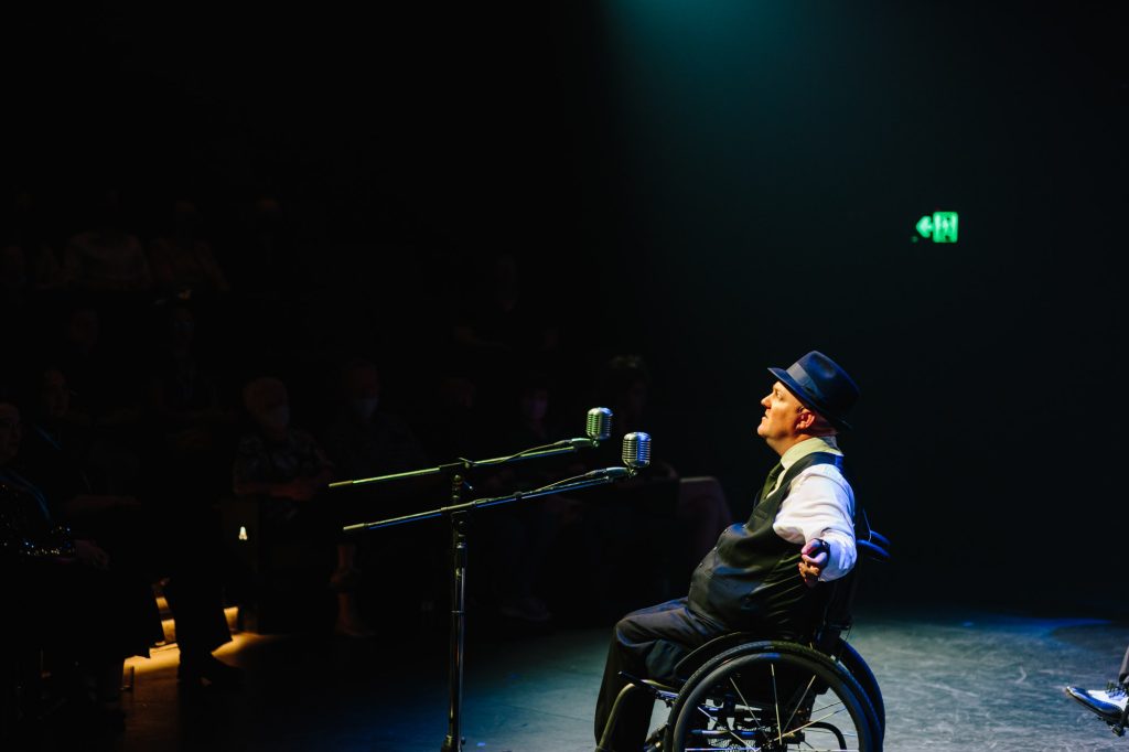 A man in a wheelchair is on stage performing. He is wearing a fedora and suit with an older styled microphone stand in front of him. He is in the middle of singing a high long note with his arms outstretched. Stage lights shine down on him.