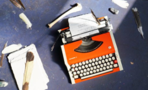 An orange old-fashioned typewriter is shot on a gray concrete floor with various white feathers laying around it. A small piece of paper is in the typewriter and there is a big pile of the same-sized paper on the left hand side.