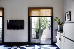 Spicers retreat room. There is a door with French doors, and a brown curtain a quarter of the way down. There is a tv mounted to the wall and white cabinets on the right side against the wall. The floor is blue and white geometric tiles.