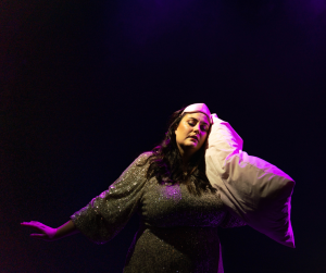 Emma-June Curik is on stage wearing a sparkly dress and a silk sleeping mask. She is mid-performance where she is holding a white pillow and has her eyes closed. Pink stage lights shine down on her and the background is completely black.