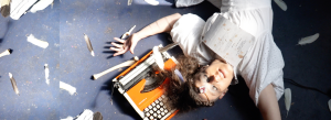 Andy is laying on the ground in a white dress near an old-fashioned orange typewriter. There are various bird feathers around. The photo is taken as an overhead shot straight down.