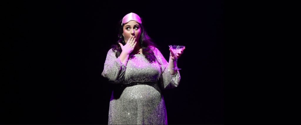 Emma-June Curik is standing and yawning. She is wearing a sparkly dress and a silk sleeping mask and is holding a filled martini glass. Pink stage lights shine down on her and the background is completely black.