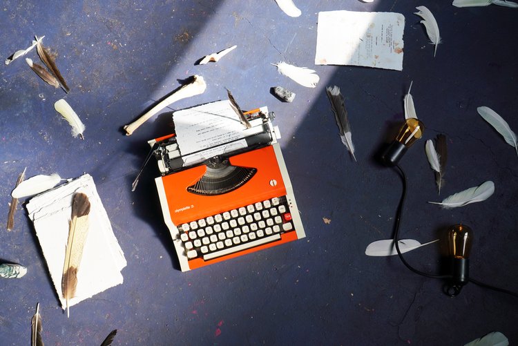 An orange old-fashioned typewriter is shot on a gray concrete floor with various white feathers laying around it. A small piece of paper is in the typewriter and there is a big pile of the same-sized paper on the left hand side.