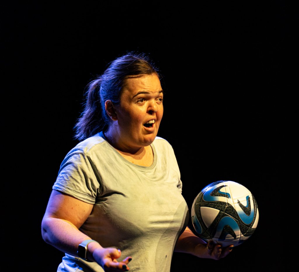 Mags captured performing against a black backdrop. Mags is holding a FIFA WWC soccer ball and appears to be mid-speech.