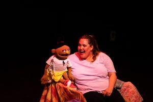 A lady wearing a pink short on stage, holding a puppet.