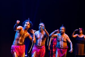 A group of indigenous performers on stage performing. They wear red shorts with white body paint on them.