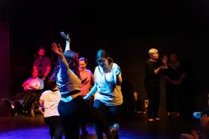 A group of people dancing on stage. Some have their arms up in the air and they are smiling.