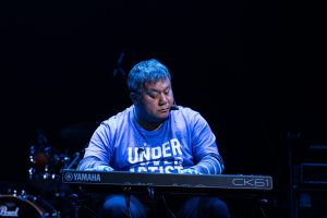 A man playing a keyboard. He is wearing a blue long sleeve shirt with the wording ' Undercover Artist' on the front.