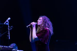 A female standing on stage performing. She is holding a microphone and has a purple light on her.