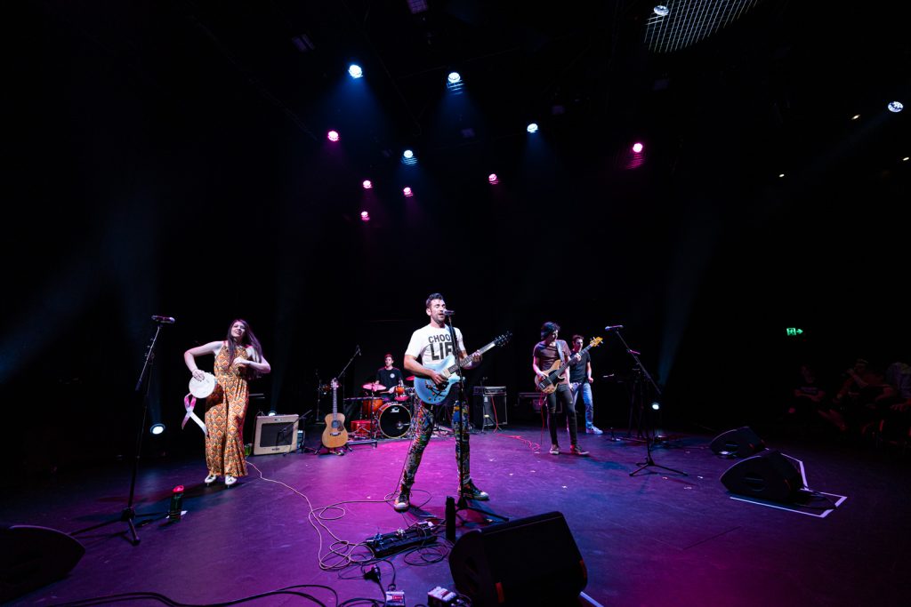 Group of musicians on stage performing with a purple stage light above them. They are all playing different instruments.