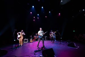 Group of musicians on stage performing with a purple stage light above them. They are all playing different instruments.