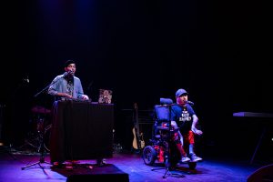 Two guys performing on stage. One is standing in front of a key board, the other in a wheelchair with a microphone in front of him.