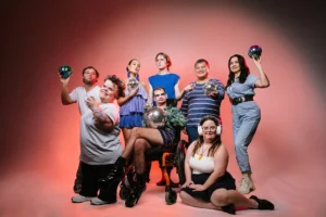 A group of people are standing in front of a white backdrop with dramatic red lights. They are all posing and a few people are holding mirror balls.