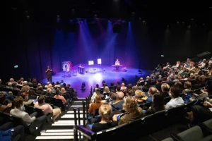 A theatre room filled with an audience where a stage is lit up by bright purple and blue lights. Under the lights are various stage props, a person playing a keyboard and another person Auslan interpreting.