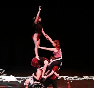 Five circus theatre performers perform an acrobatic tower the height of three people, standing and leaning on one another. One performer at the bottom of the tower uses a wheelchair.