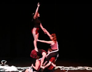 Five circus theatre performers perform an acrobatic tower the height of three people, standing and leaning on one another. One performer at the bottom of the tower uses a wheelchair.