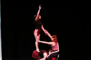 Circus performers create a human pyramid on stage against a black background. One performer balances on top with an arm raised high, while the others support from below, showcasing strength, trust, and teamwork in motion.