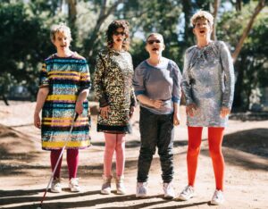 4 Individuals standing outside on a dirt pathway with trees behind them. They are wearing glittery outfits, one with a white cane. They are posing for the camera.