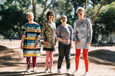 4 Individuals standing outside on a dirt pathway with trees behind them. They are wearing glittery outfits, one with a white cane. They are posing for the camera.