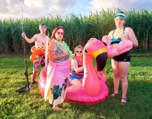 A group of people standing in a together with sugar cane crop behind them. They are wearing swimming costumes, and holding different inflatable swimming toys.