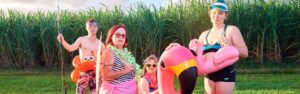 A group of people standing in a together with sugar cane crop behind them. They are wearing swimming costumes, and holding different inflatable swimming toys.