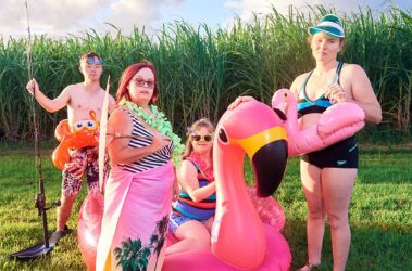 A group of people standing in a together with sugar cane crop behind them. They are wearing swimming costumes, and holding different inflatable swimming toys.