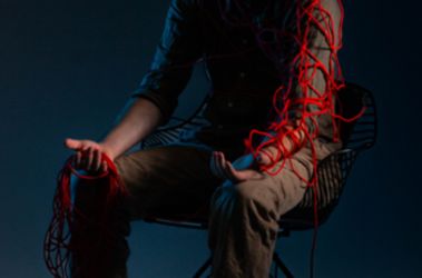 A person sits in a wireframe chair under moody, low lighting. They are partially wrapped in tangled red yarn, which drapes over their shoulders and arms.