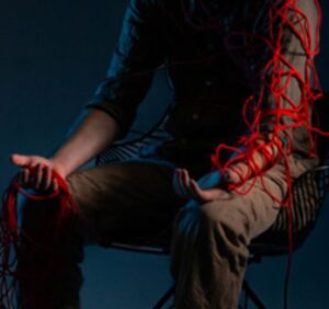 A person sits in a wireframe chair under moody, low lighting. They are partially wrapped in tangled red yarn, which drapes over their shoulders and arms.