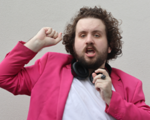 Man wearing a bright pink jacket, with a white t-shirt underneath. He has headphones around his neck and his arms are up