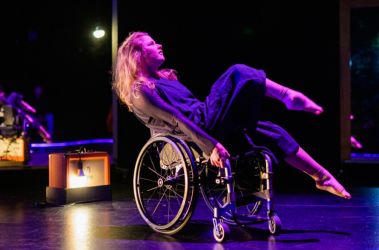A performer on stage in a wheelchair, balancing on her back two wheels, with the two front off the ground. Her gaze is forward, and she has red and purple stage lights on her.