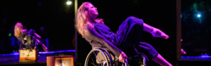 A performer on stage in a wheelchair, balancing on her back two wheels, with the two front off the ground. Her gaze is forward, and she has red and purple stage lights on her.