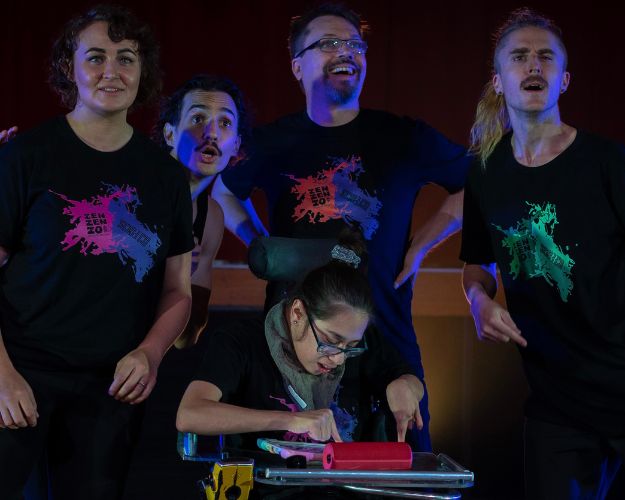 A dynamic performance scene featuring 5 performers on stage. One performer is in a wheelchair using an ipad while four dancers around her strike expressive poses, captured mid-motion with intense focus and energy. They all wear black outfits with colourful abstract designs. The stage is lit with dramatic purple and pink lighting.