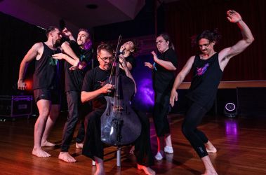 A dynamic performance scene featuring six performers on stage. One musician plays an electric cello while five dancers around him strike expressive poses, captured mid-motion with intense focus and energy. They all wear black outfits with colourful abstract designs. The stage is lit with dramatic purple and pink lighting.